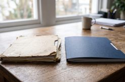 Two contract folders sitting side by side on an office desk, one visibly older and worn than the other, soft daylight...